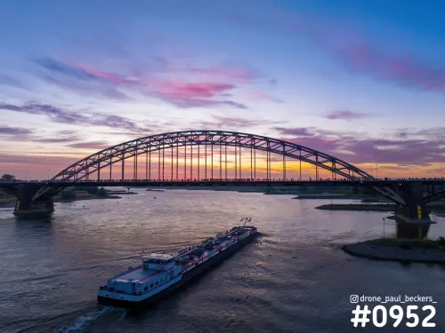 Luchtfoto van de Waalbrug in Nijmegen bij zonsondergang, met een vrachtschip op de Waal en mensen op het brugdek