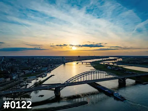 Luchtfoto van de Waalbrug in Nijmegen bij zonsondergang, met een binnenvaartschip op de Waal en de Oversteek in de verte