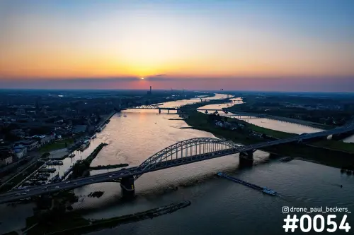 Luchtfoto van de Waalbrug in Nijmegen bij zonsondergang, met de Waalkade links en De Oversteek ver op de Waal