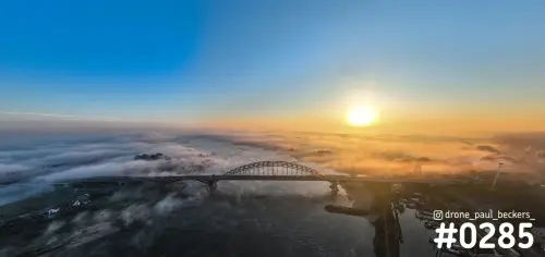 Panorama-luchtfoto van de Waalbrug in Nijmegen bij zonsopkomst, met mistbanken boven de Waal en de Ooijpolder