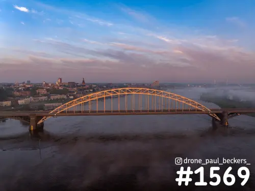 Luchtfoto van de Waalbrug in Nijmegen bij zonsopkomst, de stalen boog in oranje licht en mist boven de Waal