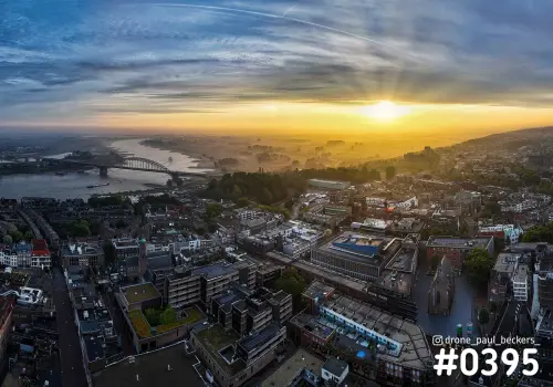 Luchtfoto van Nijmegen bij zonsopkomst met de Waalbrug, de Waal en mist boven de uiterwaarden achter de daken