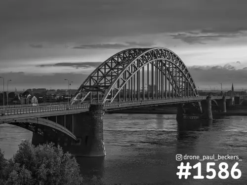 Zwart-wit luchtfoto van de Waalbrug in Nijmegen met de stalen boogconstructie boven de rivier de Waal, onder een bewolkte lucht