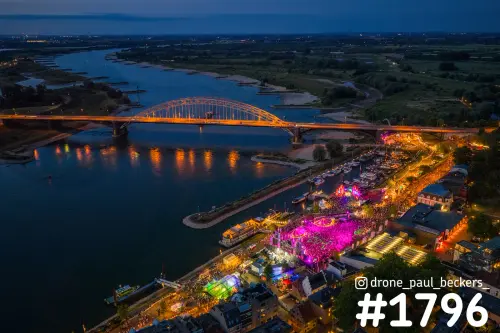 Luchtfoto van de Waalbrug en de Waalkade in Nijmegen in de avond, met roze verlicht festivalterrein aan de Waal