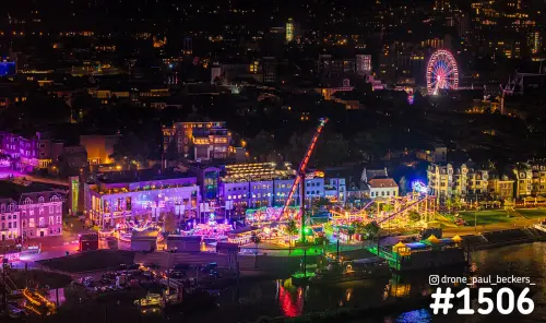 Luchtfoto van de kermis op de Waalkade in Nijmegen bij nacht, met verlicht reuzenrad en kermisattracties aan de rivier