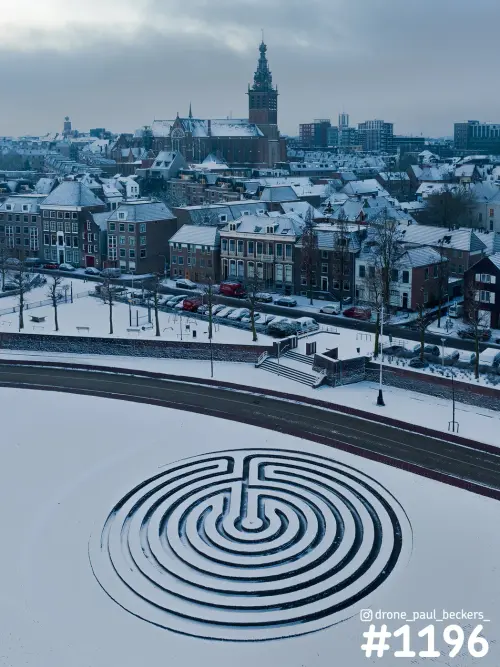 Luchtfoto van de Waalkade in Nijmegen met sneeuw, een labyrintpatroon op het plein en de Stevenskerk op de achtergrond