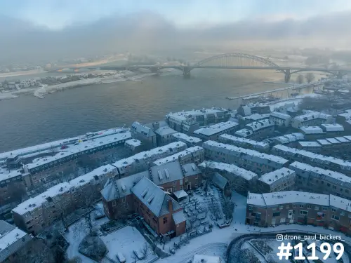 Luchtfoto van besneeuwde daken aan de Waalkade in Nijmegen, met de Waalbrug in de mist boven de rivier de Waal