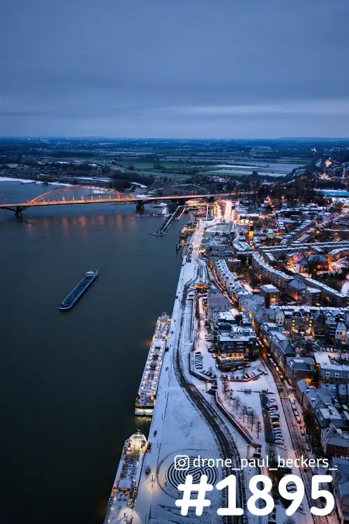 Luchtfoto van de Waalkade in Nijmegen in de schemering, sneeuw op de kade en daken, een binnenvaartschip op de Waal bij de verlichte Waalbrug