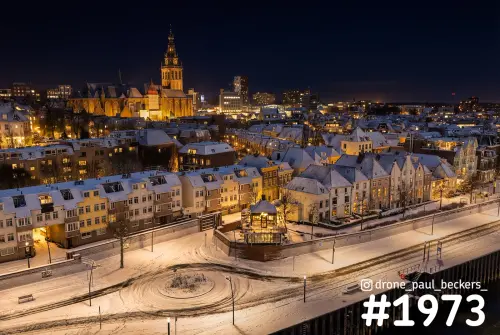Luchtfoto van de Waalkade in Nijmegen in de avond onder sneeuw, met de verlichte Stevenskerk boven besneeuwde daken