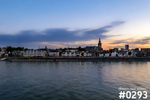 Nijmegen met de Stevenskerk en de Waalkade vanaf de Waal in de schemering, met oranje gloed en een brede wolkenband