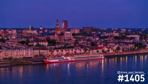 Luchtfoto van de Waalkade in Nijmegen in het blauwe uur, met een rivierschip aan de kade en de verlichte Stevenskerk