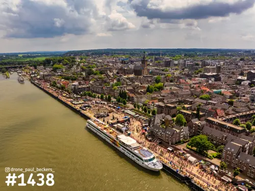 Luchtfoto van de Waalkade in Nijmegen met een riviercruiseschip aan de kade, drukke menigte en de Stevenskerk op de achtergrond