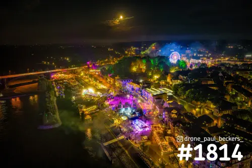 Luchtfoto van de Waalkade in Nijmegen bij nacht, met paars verlichte feesttenten, een reuzenrad en de maan boven de Waal