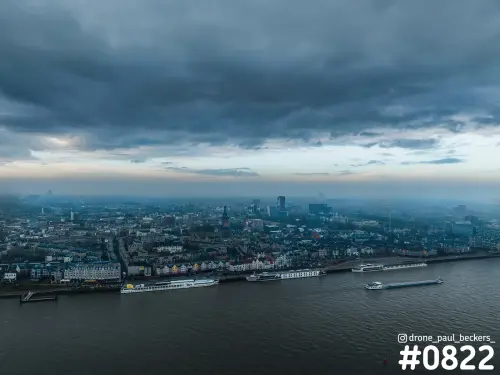 Luchtfoto van de Waalkade en de Waal in Nijmegen onder zware wolken, met schepen op de rivier en mist boven de stad