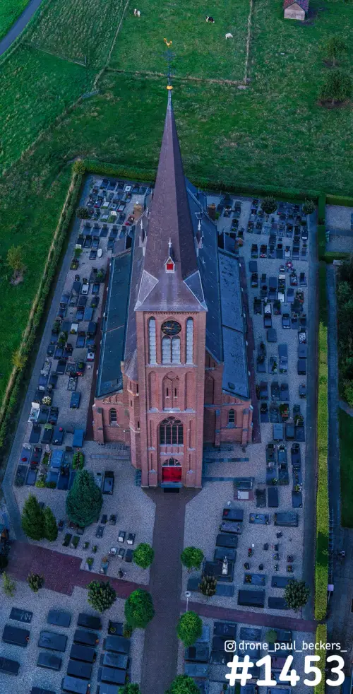 Luchtfoto van een bakstenen kerk met hoge toren in Weurt, recht van boven, omringd door het kerkhof en groene weilanden