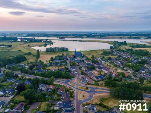 Luchtfoto van Weurt bij Nijmegen in de schemering, met kerktoren, woonwijk en een waterplas in de uiterwaarden