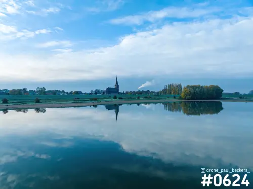 Luchtfoto van Weurt bij Nijmegen: kerktoren weerspiegeld in stil water, met herfstbomen en een witte rookpluim