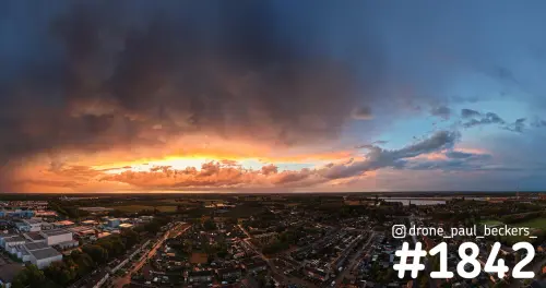 Luchtfoto van Weurt bij avondlicht, met donkere wolken en een oranje gloed boven woonwijk en bedrijventerrein
