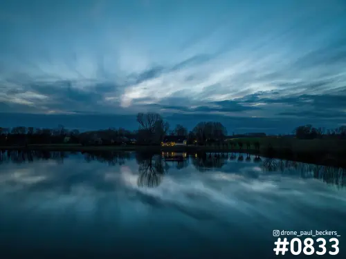 Luchtfoto van Weurt in het blauwe schemerlicht, met een verlicht huis en kale bomen weerspiegeld in stil water