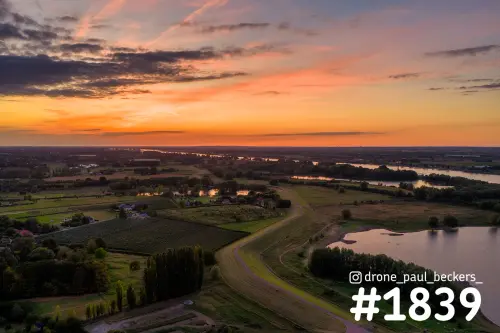 Luchtfoto van de uiterwaarden bij Weurt tijdens zonsondergang, met een dijk, akkers en water onder een oranje wolkenlucht