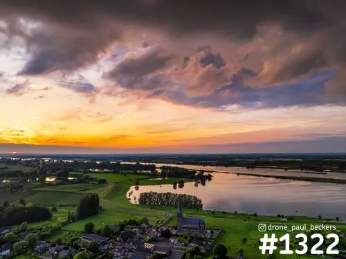 Luchtfoto van Weurt aan de Waal bij zonsondergang, met kerktoren, water in de uiterwaarden en zware wolken