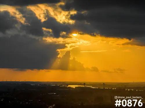 Luchtfoto bij Weurt met zonnestralen door donkere wolken, oranje lucht en een rivier met brug aan de horizon