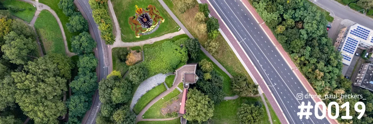 Luchtfoto van het Valkhof gebied in Nijmegen met een bloemperk in wapenvorm, een brede weg met fietspad en zomergroene bomen