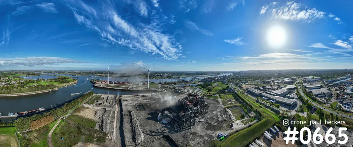 Luchtpanorama van de Waal en een industrieterrein met sloopwerk, stof boven de staalconstructie en windturbines in de verte