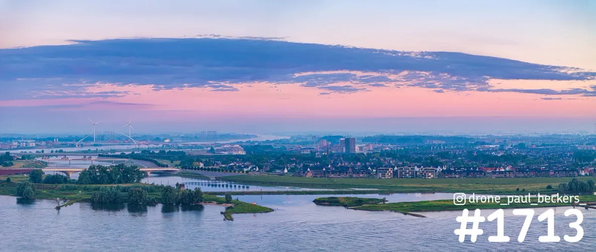 Panorama-luchtfoto van de Waal bij Nijmegen-Lent met de Oversteek op de achtergrond, onder een roze avondlucht met brede wolkenband