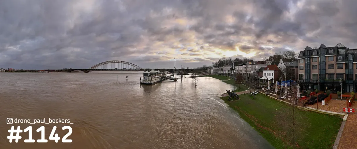 Panorama van de Waal in Nijmegen met hoog water, de Waalbrug in de verte en de Waalkade onder een dreigende wolkenlucht