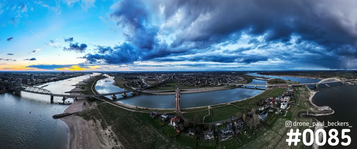 Panorama-luchtfoto van de Waal bij Nijmegen en Lent met Waalbrug en De Oversteek onder een dreigende blauwe wolkenlucht