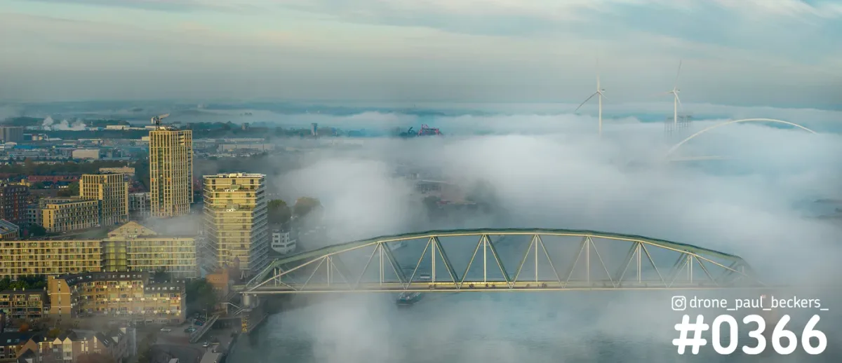 Luchtfoto van Nijmegen met mist boven de Waal, een stalen vakwerkbrug en de boog van De Oversteek op de achtergrond