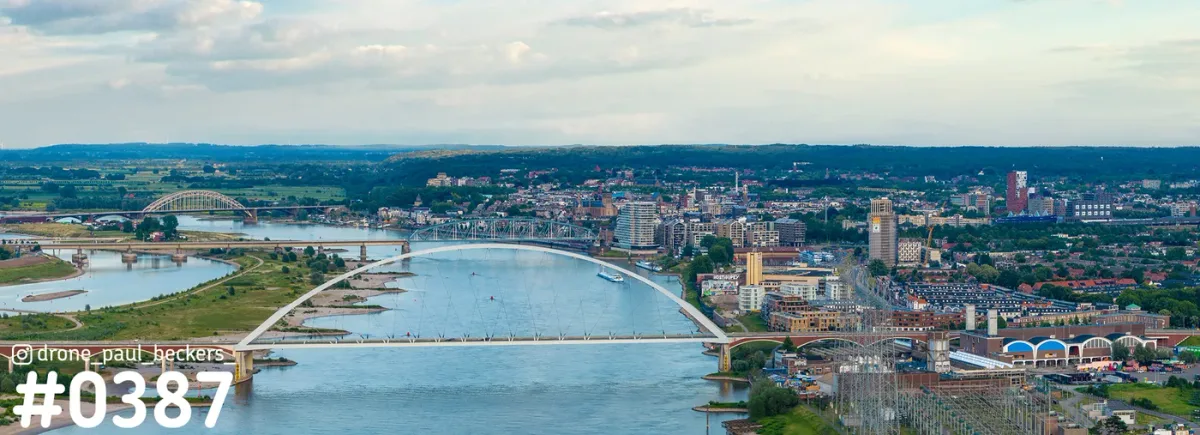 Panorama van de Waal in Nijmegen met een witte boogbrug op de voorgrond en de Waalbrug in de verte onder bewolkte lucht