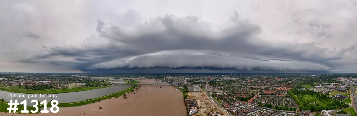 Panoramafoto van een brede shelf cloud boven de Waal en Nijmegen, met bruin rivierwater en de nevengeul bij Lent links