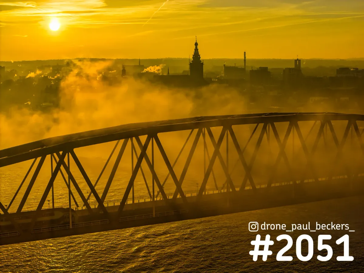 Luchtfoto van de stalen boogbrug over de Waal in Nijmegen bij zonsopkomst, met mist en de toren van de Stevenskerk