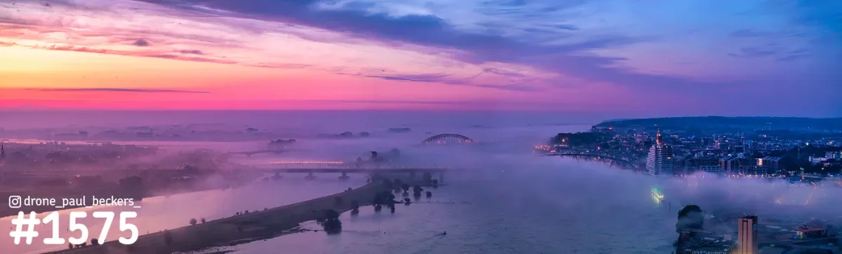 Luchtfoto van de Waal bij Nijmegen met Waalbrug in de verte, roze ochtendlucht en mist die over de Handelskade trekt
