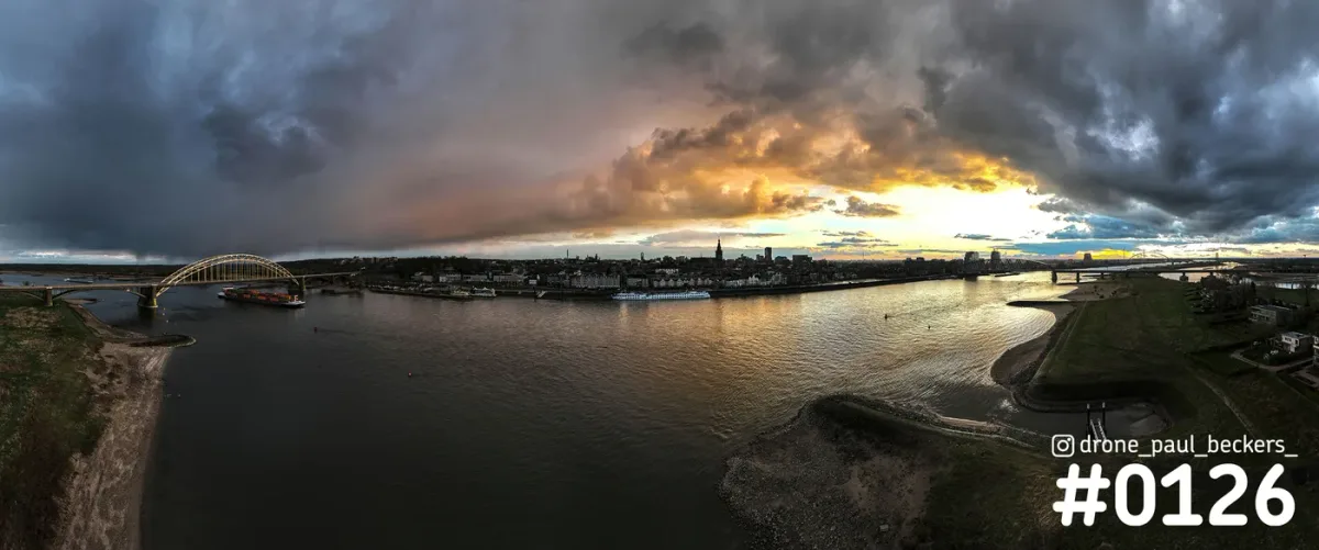 Panorama van de Waal in Nijmegen met Waalbrug en Stevenskerk onder dreigende wolken en oranje avondlicht