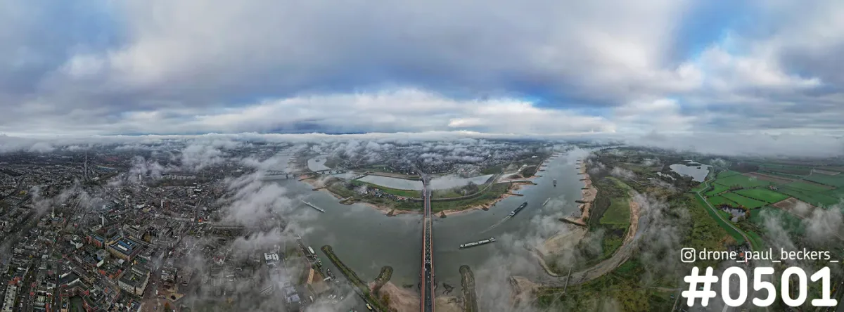 Panorama-luchtfoto van de Waal en de Waalbrug in Nijmegen, met lage mist boven de stad en een zwaar bewolkte lucht