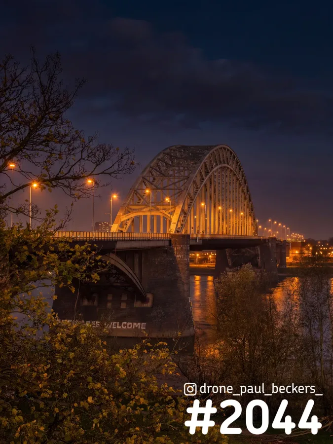 Waalbrug in Nijmegen in de avondschemering, met verlichte boog, oranje lantaarns en weerspiegeling in de Waal