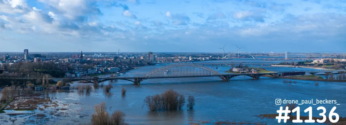 Panorama van de Waalbrug in Nijmegen bij hoogwater, met overstroomde uiterwaarden en De Oversteek op de achtergrond