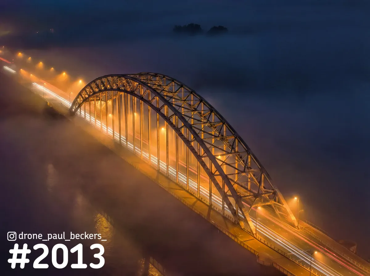 Luchtfoto van de Waalbrug in Nijmegen in de avondmist, met oranje verlichting en lichtstrepen van het verkeer