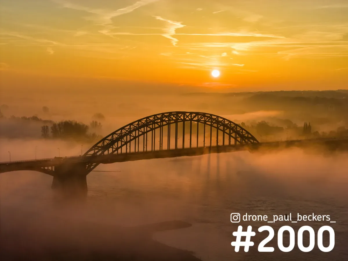 Luchtfoto van de Waalbrug in Nijmegen bij zonsopkomst, met laagstaande zon en oranje mist boven de rivier de Waal