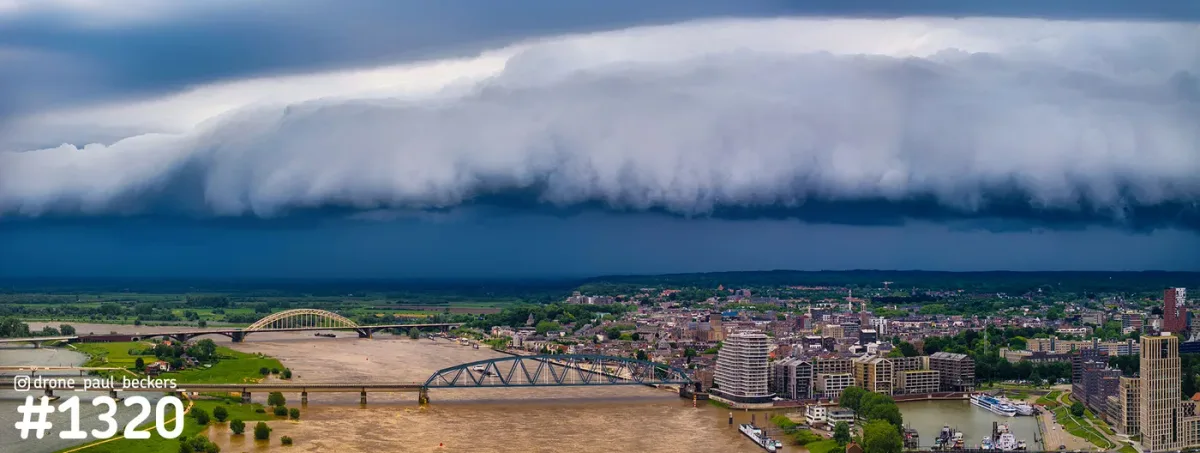 Luchtfoto van een brede onweerswolk boven de Waal en de Waalbrug in Nijmegen, met de stad en Lent eronder