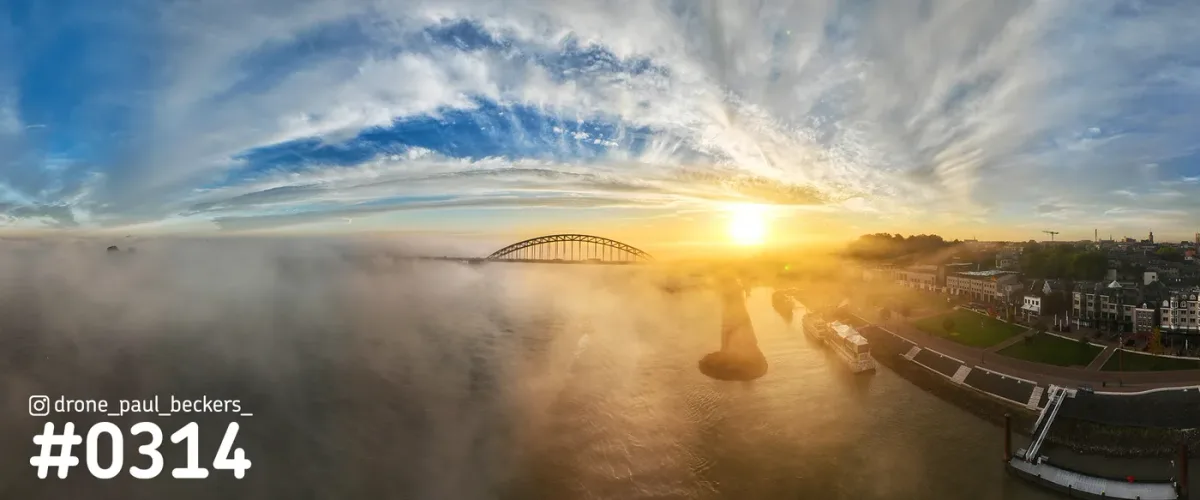 Panoramafoto van de Waalbrug in Nijmegen bij zonsopkomst, met mist boven de Waal en een schip langs de Waalkade