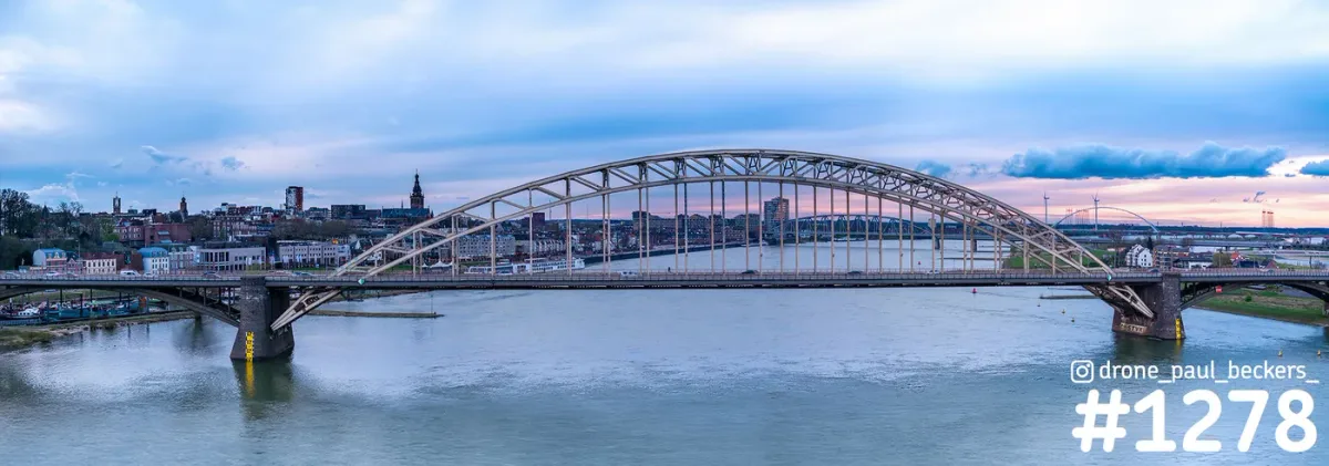 Panoramafoto van de Waalbrug in Nijmegen met de Stevenskerk links erachter, onder een roze en blauwe schemerlucht