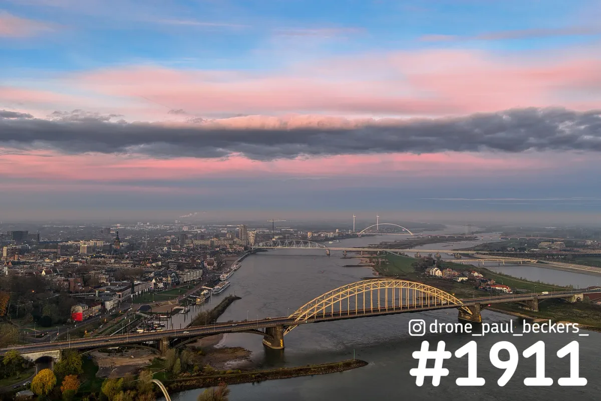 Luchtfoto van de Waalbrug in Nijmegen bij schemering, met roze wolken boven de Waal en De Oversteek in de verte