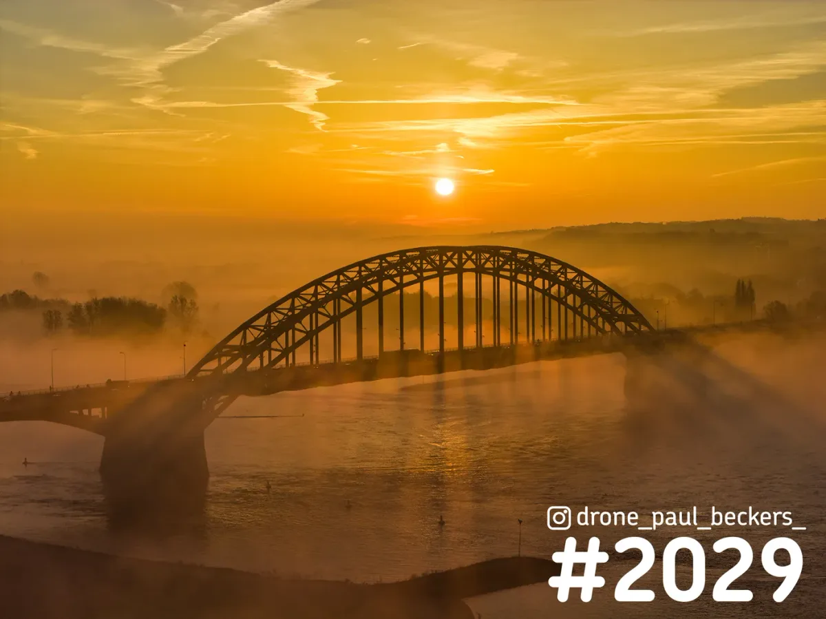 Luchtfoto van de Waalbrug in Nijmegen bij zonsopkomst, met de zon boven de boogbrug en mist die over de Waal trekt