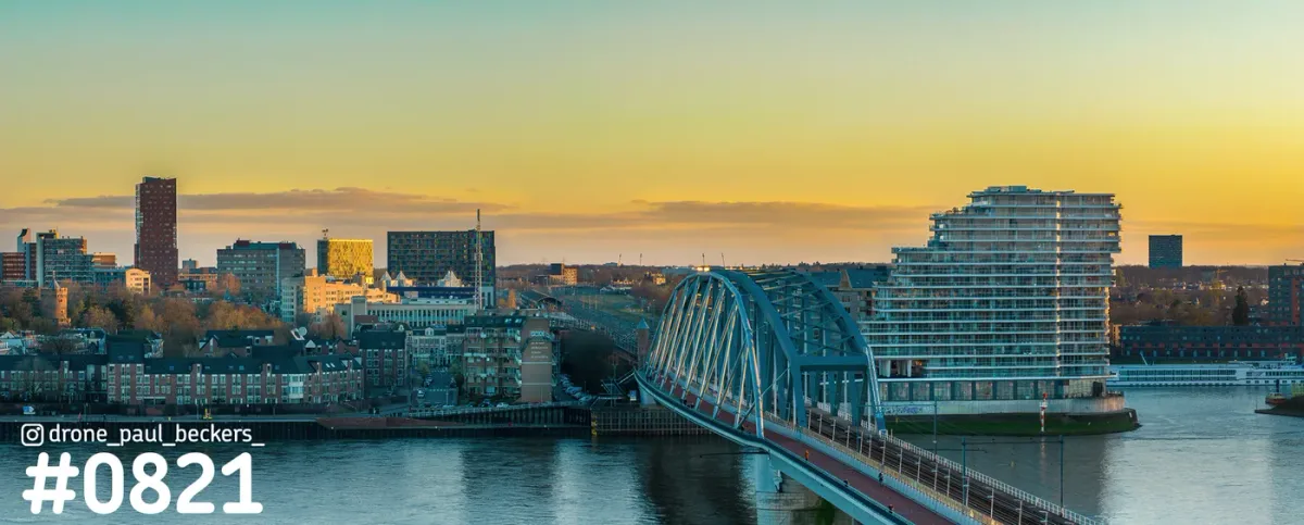 Panorama van de Waalkade in Nijmegen met het Nimbus-gebouw en een blauwe boogbrug over de Waal in het gouden uur